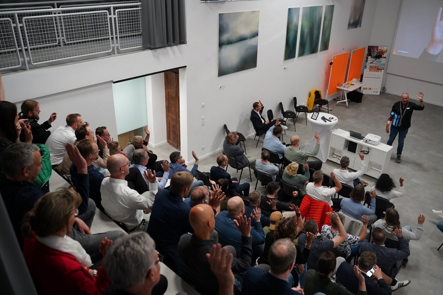 Ein großes Publikum sitzt in einer modernen Eventlocation und hört einem Vortrag zu.