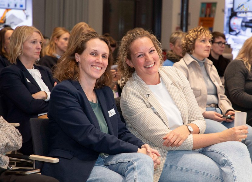 Zwei Frauen sitzen in einem Publikum und lächeln in die Kamera.