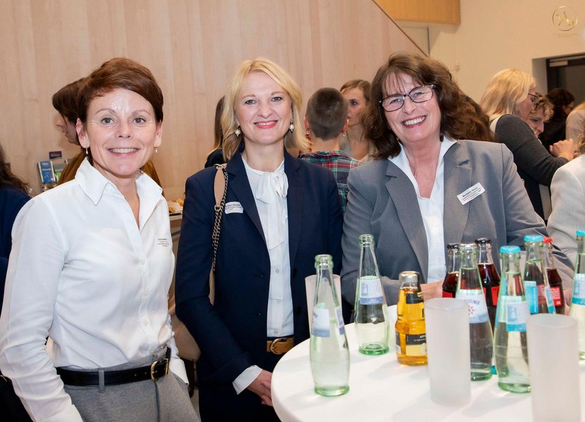 Drei Frauen stehen an einem Stehtisch und lächeln in die Kamera.