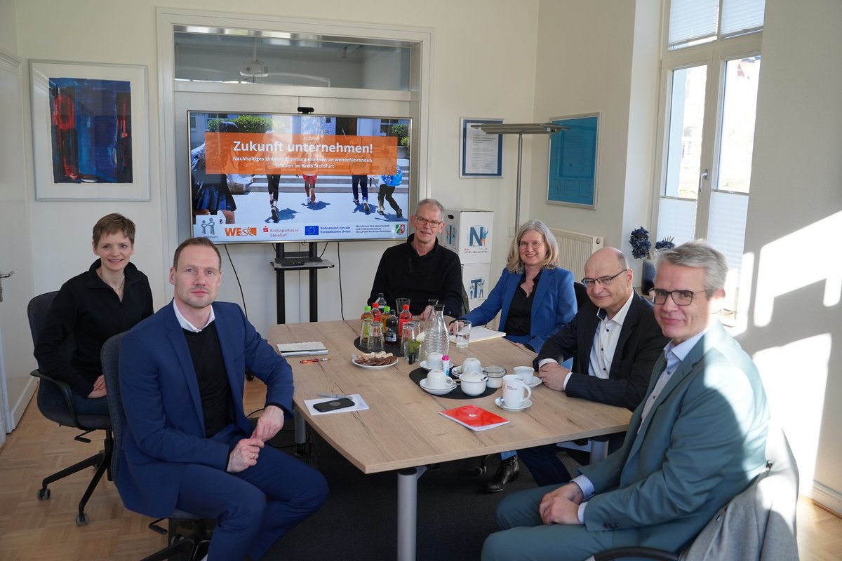 Sechs Personen sitzen um einen Besprechungstisch und schauen für das Foto in die Kamera. Im Hintergrund steht ein Monitor auf dem der Text "Zukunft unternehmen!" steht.